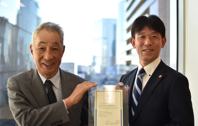 ＜認証書授与式の様子(左：UCDA理事長　在間 稔允 様、右：当社執行役員　下間 博仁)＞